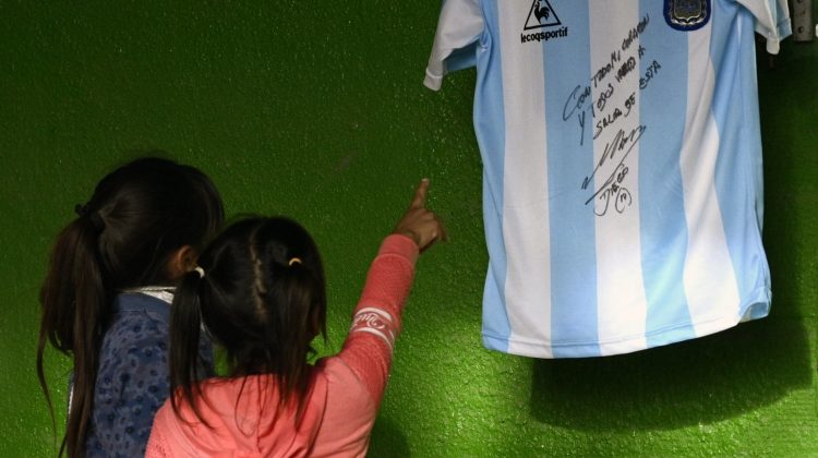 Kids watch a replica of Argentina's football team jersey used during the final of the FIFA World Cup Mexico '86, handwritten and signed by Argentine former football star Diego Maradona hanging from a Catholic altar while they wait for the afternoon milk at a community kitchen during the lockdown imposed by the government against the spread of the new coronavirus, COVID-19, in Jose C. Paz, Buenos Aires, Argentina, on May 8, 2020. - Maradona's signed jersey and reading With all my heart and we are all going to get over this will be raffled among those who donate food for people at a working class neighbourhood called Rene Favaloro, called after the Argentine cardiac surgeon. (Photo by JUAN MABROMATA / AFP)